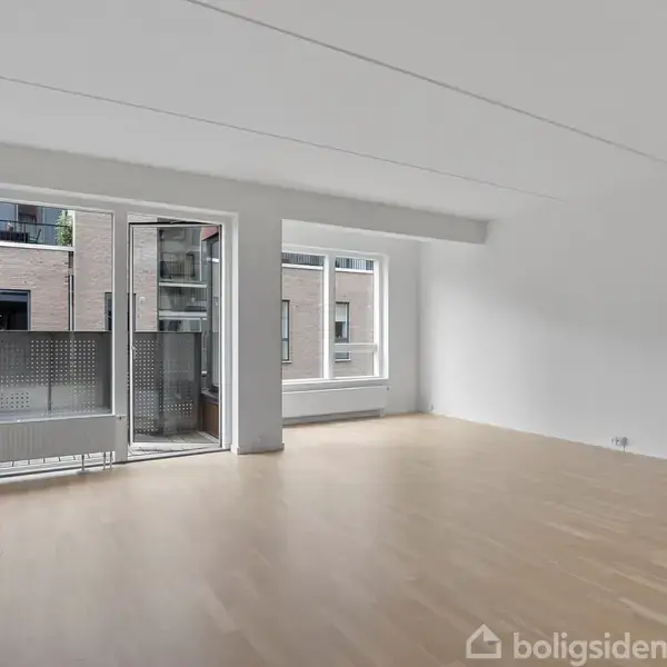 Et tomt rum med trægulv, hvor en dør og store vinduer åbner mod en balkon. Rummet fremstår lyst og moderne, med udsigt til en murstensbygning udenfor.
