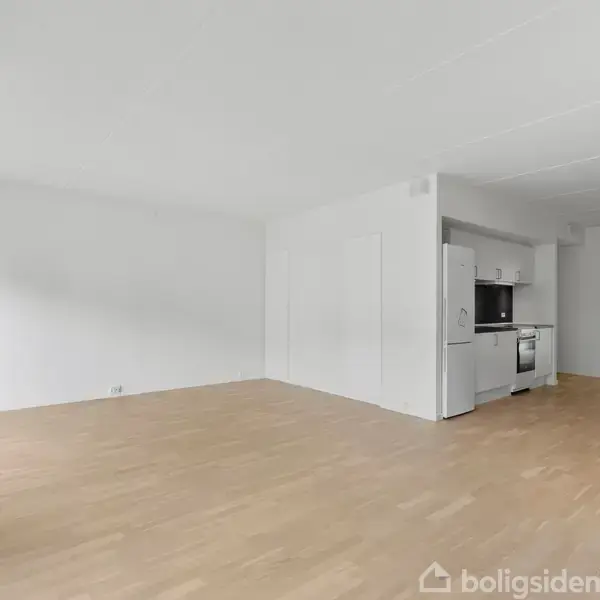 En tom stue med trægulv har en åben køkkenløsning med hvide skabe i et moderne hjem. Lyse vægge og loft skaber et minimalistisk udtryk i et rummeligt interiør.