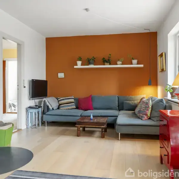 Stue med blå sofa placeret mod en orange væg, dekoreret med hylde og planter. Rød kommode står ved siden af et vindue. Lampe og musikstander giver lys og atmosfære.