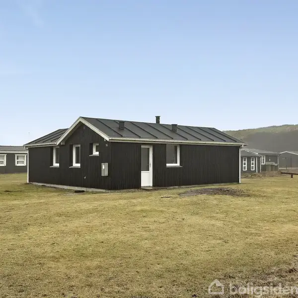 Et sort træhus står på en græsplæne. I baggrunden ses flere mindre huse og en bakket landskab under en klar himmel.