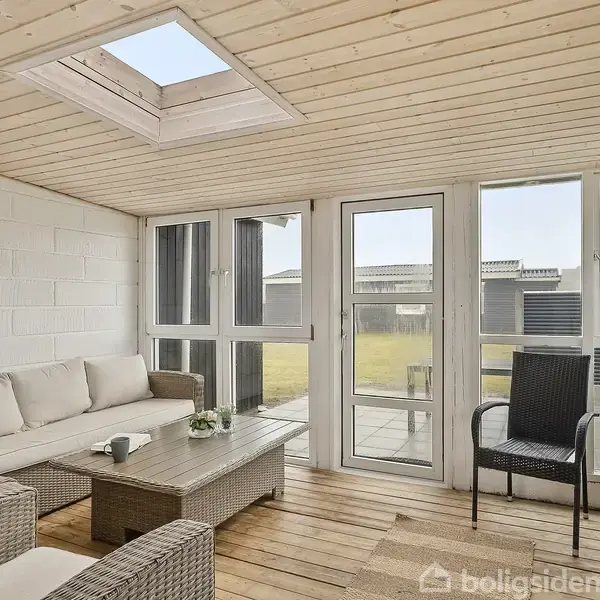 En lys vinterhave med træloft og glasdør, indeholder sofa, lænestol og bord. Udsigt til en have udenfor. Stemningen er indbydende med afslappende møbler i flettet design.