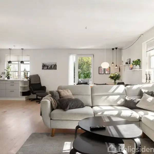Sofa i lys stue fungerer som rumdeler mod køkkenet. Stort vindue og dør fører til have. Modern belysning og grønne planter skaber en hyggelig atmosfære.