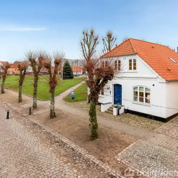 Hvidt hus med rødt tag står ved en brostensbelagt gade. I forgrunden er træer uden blade. I baggrunden ses flere huse og en græsbevokset bakke.