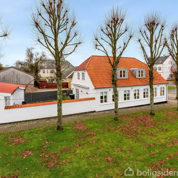 Hus med rødt tegltag ligger bag en hvid mur, omgivet af græsplæne og træer i en bymæssig kontekst. En bred indkørsel ses ved siden af huset.