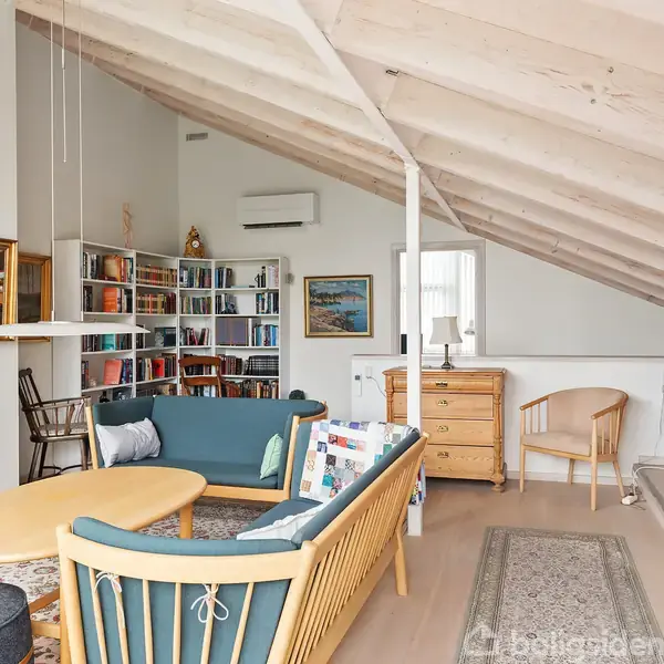 Loftstue med skrå loft, indeholder en blå sofa ved et ovalt bord, reoler fyldt med bøger, samt kommode og stole. Indrammede malerier dekorerer væggene. Lys og hyggelig atmosfære.