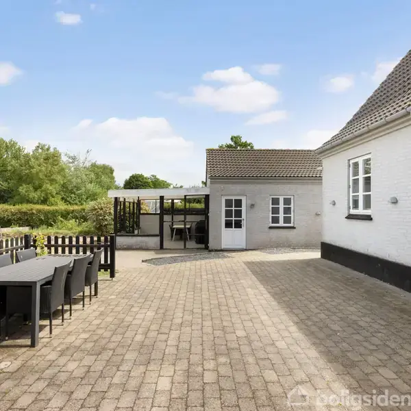 Et udendørs spisebord med stole står på en brolagt terrasse ved siden af et hvidt hus. Stolper og hegn omkranser området, og en græsplæne med legeredskaber ses i baggrunden.