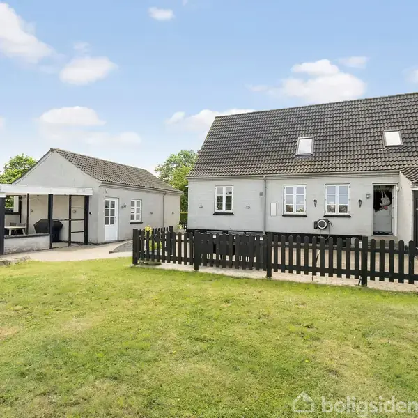 Et hvidt hus med skråt tag står i en have med græsplæne. Til venstre er en åben carport. Hegn omkranser området.