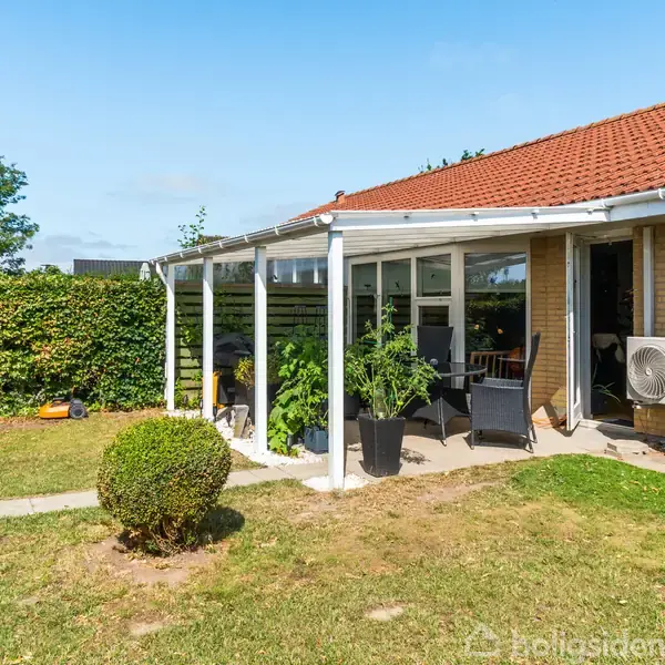 En terrasse med havemøbler under en hvid pergola. Murstenshus med rødt tegltag i en solrig have, omgivet af grønne buske og en veltrimmet græsplæne.