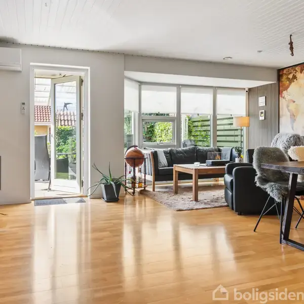 Stue med sofa og sofabord i centrum, placeret på et tæppe. Spisestue i baggrunden, globus ved siden af sofaen. Lys fra store vinduer. Åben dør mod terrasse.