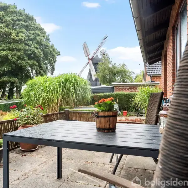 Et bord med en blomsterpotte står på en terrasse. En vindmølle ses i det frodige landskab med store træer og buske, ved siden af et murstenshus.