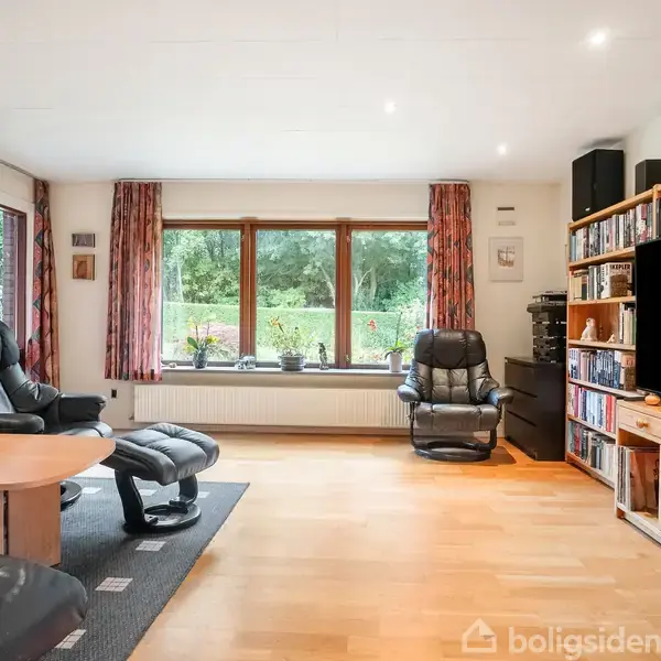 En hyggelig dagligstue med sorte lænestole og et stort fladskærms-TV placeret på en reol fyldt med bøger. Store vinduer med udsigt til en grøn have.