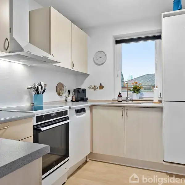 Et moderne køkken med hvide skabe og sorte bordplader. Der står en ovn, en emhætte og et køleskab. Vinduet viser udsigt. Tekst: "Boligsiden". En vægur hænger på væggen.