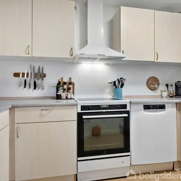 Et køkken med lyse skabe indeholder en ovn i midten, omgivet af en opvaskemaskine og køkkenborde. På væggen er der en knivmagnet, krydderiglas og et ur.