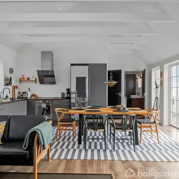 Sofa med puder i moderne stue-køkken alrum, spisebord med stole på tæppe. Store vinduer lader lys komme ind; minimalistisk køkken langs én væg. Mørke trægulve, hvide vægge.