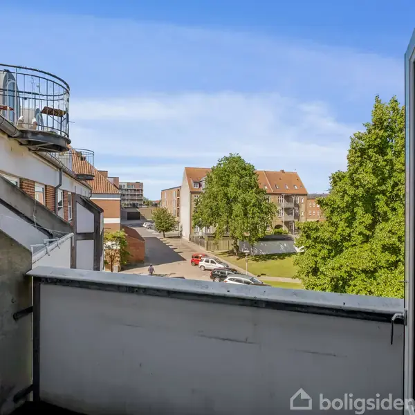 En lille altan med et gelænder, der vender ud mod en gade. I baggrunden ses lejlighedsbygninger, træer og en park, oplyst af dagslys.