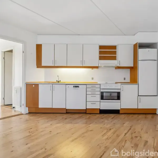 Et køkken med hvide skabe og trædetaljer står mod en hvid væg. Køleskab, ovn og opvaskemaskine er integreret. Trægulv strækker sig ud i rummet.