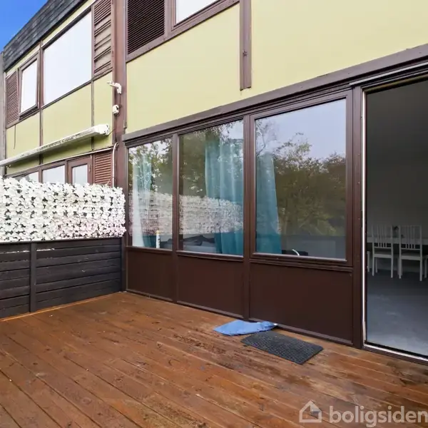 Terrasse med trædæk foran hus med store glasvinduer. Blå måtte ligger ved døråbning. Indvendigt ses spisestuemøbler. Væg af blomsterdekoration til venstre, blå himmel ovenfor.