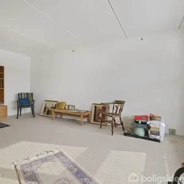 Et næsten tomt rum med en træreol på venstre væg. Et bord og stol er placeret mod højre væg. Tæpper og nogle kasser ligger på gulvet.