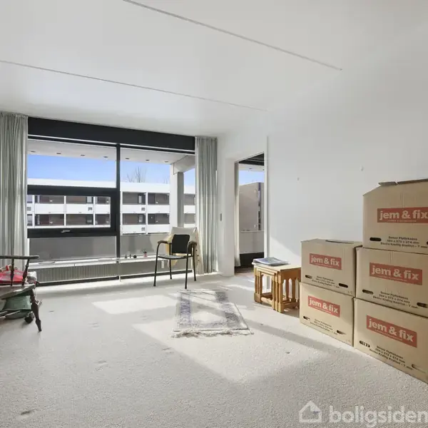 Et lyst rum med en stor vinduesfront. Papkasser mærket "jem & fix" stablet på gulvet. To stole og et lille bord står ved vinduet. Nogle malerier står op ad væggen.