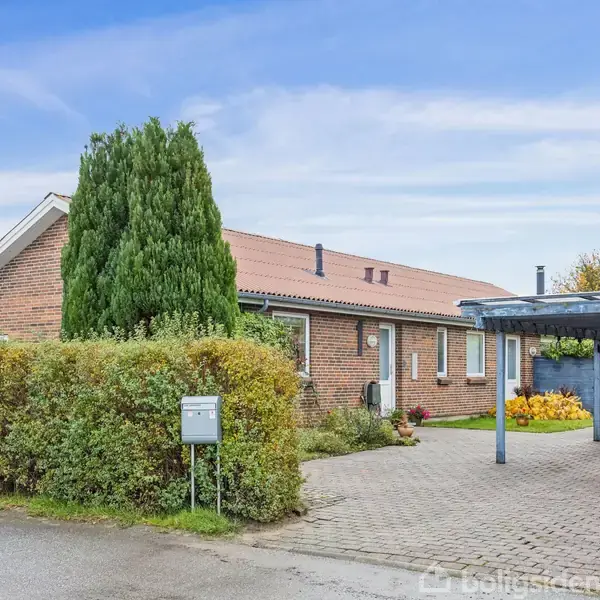 En murstensvilla med rødt tag, omkranset af en velplejet hæk, står ved siden af en carport. Haven er dekoreret med buske og en høj, tætvokset træ.