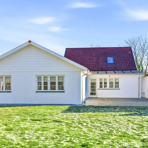 Et hvidt hus står i en have med en græsplæne, med store vinduer og et rødt tag. Klar blå himmel og træer i baggrunden.