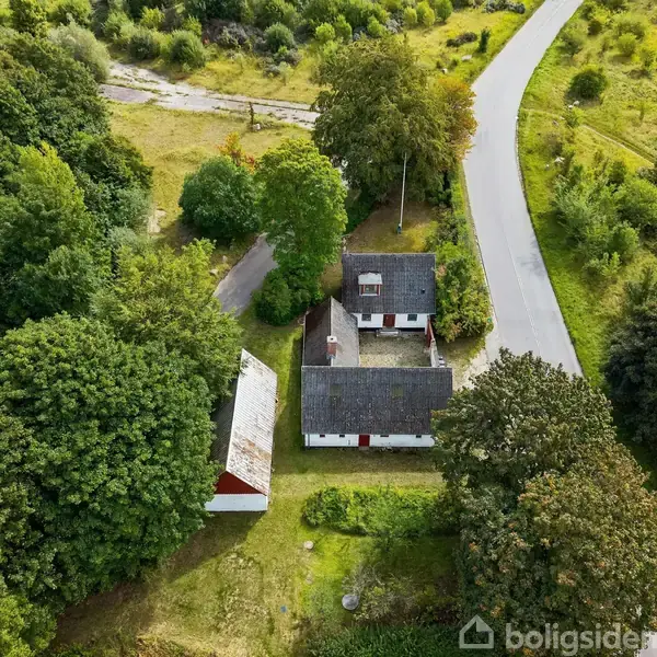 To hvidmalede bygninger står omgivet af tætte træer. En vej løber forbi bebyggelsen, der er placeret i et grønt, landligt område.