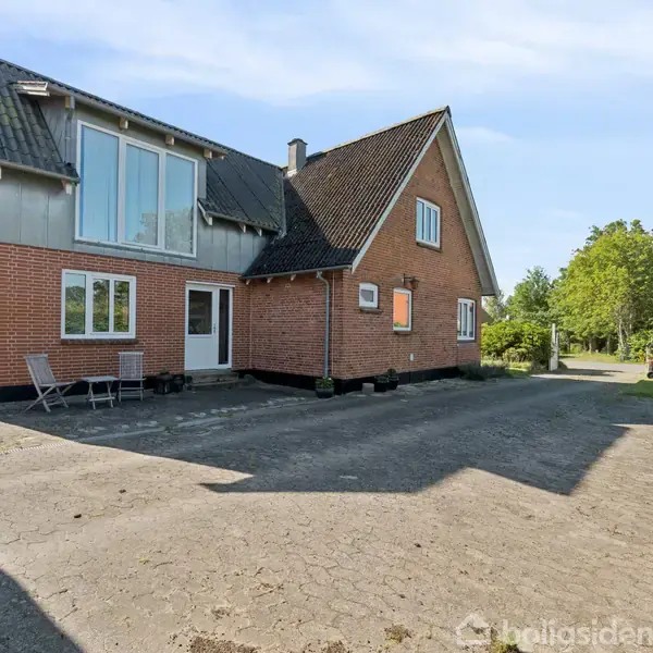 Rødt murstenshus med store vinduer og sadeltag står på en stor, asfalteret gårdsplads. To stole og et bord står foran huset. Omgivet af træer og buske.