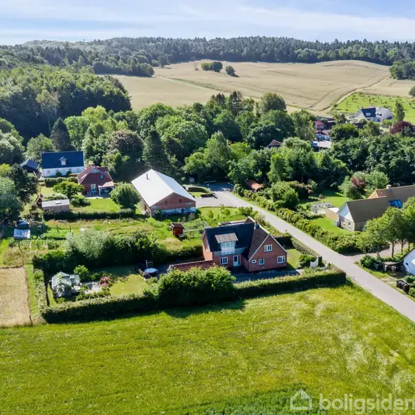 Huse ligger i en landlig landsby med veje omringet af marker og skove. Det er en lys solskinsdag med grønne træer og marker i baggrunden.