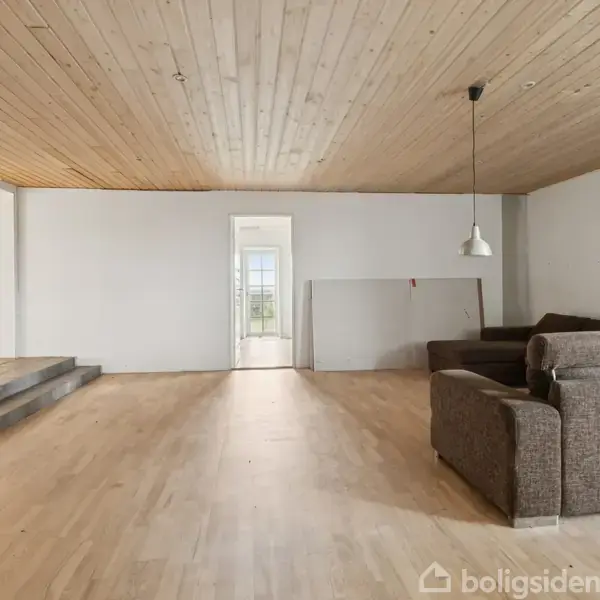 En åben stue med en brun sofa placeret til højre, træloft og trægulv, en loftslampe hængende ned, få trin til venstre fører op til en gang.