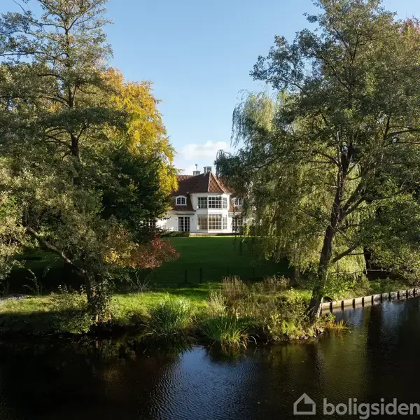 Et hvidt hus står stille i en frodig have omkranset af store træer. Foran er en sø med spejlblank overflade, der skaber en rolig atmosfære.