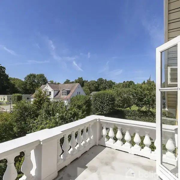 En hvidmalet balkon med åbne franske døre tilbyder udsigt over en frodig have og nabohuse i et solrigt kvarter.