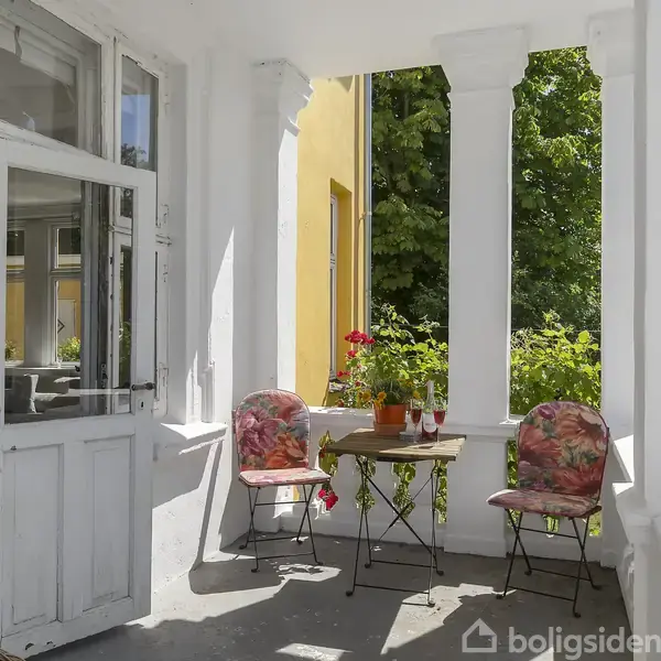 En terrasse med to stoler og et lille rundt bord med planter i krukker. Omkring er der hvide søjler og en gul væg, omgivet af grønne træer i baggrunden.