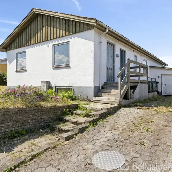 Huset står på en skrånende indkørsel med trapper, flankeret af en garage og grøn beplantning. Nummeret "2" er synligt ved hoveddøren.
