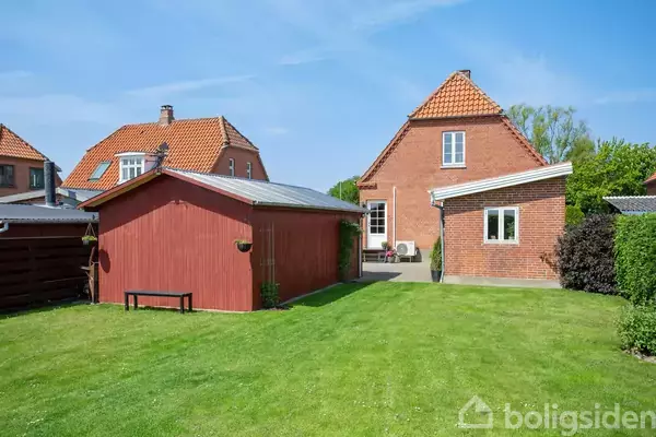 Rødt murstenshus med stejlt tag står ved siden af en rød trægarage. En velplejet græsplæne strækker sig foran bygningerne i en solrig have. Ingen tekst i billedet.