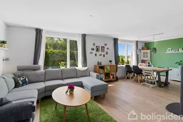 En grå hjørnesofa står i en lys stue med store vinduer. Et rundt sofabord og grønne planter tilføjer farve. Et spisebord med stole ses i baggrunden ved en grøn væg.