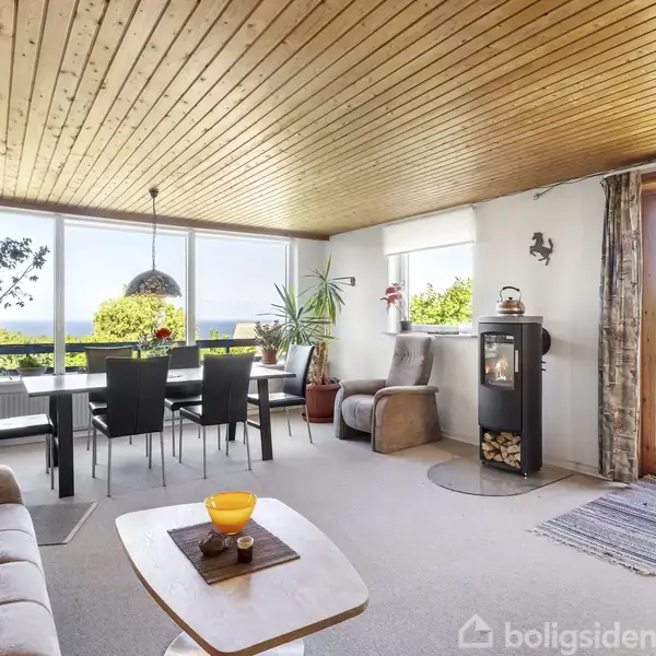 Stue med sofa, bord, spisebord med stole og brændeovn. Store vinduer og glasdør giver udsigt til træer og havet, der skaber en lys og åben atmosfære.