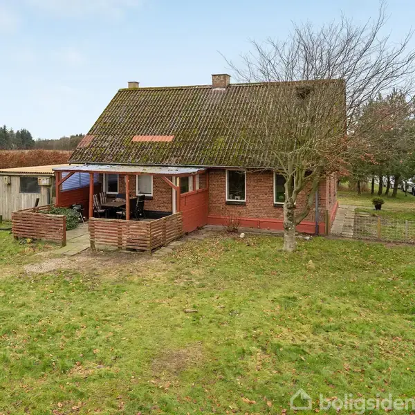 Rødt hus med skråtag udenfor, omgivet af græsplæne og buske. Foran huset ses en terrasse med tag. En stor træ står ved siden af bygningen.