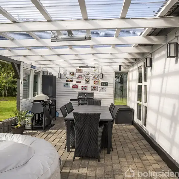 Et overdækket terrasseområde med et bord og seks stole i midten. Til venstre står en grill og nogle planter. Baggrunden viser en have med græs og træer.