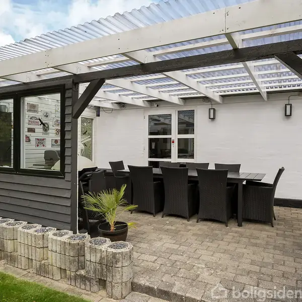 Udendørs terrasse med spisebord og sorte stole står under en pergola med gennemsigtigt tag. Omgivet af en græsplæne og op ad en grå og hvid bygning. Ingen tekst fundet.