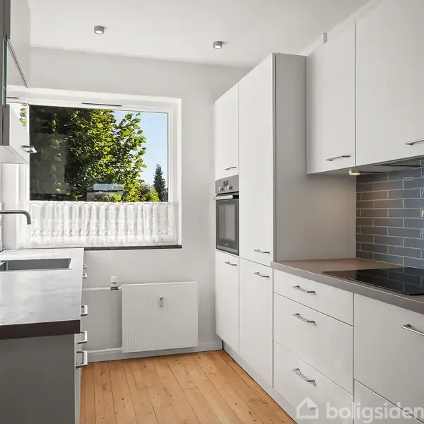 Et moderne køkken har hvide skabe, indbyggede apparater og en mørk bordplade. Et vindue giver udsigt til træer udenfor, mens et trægulv fuldender rummet.