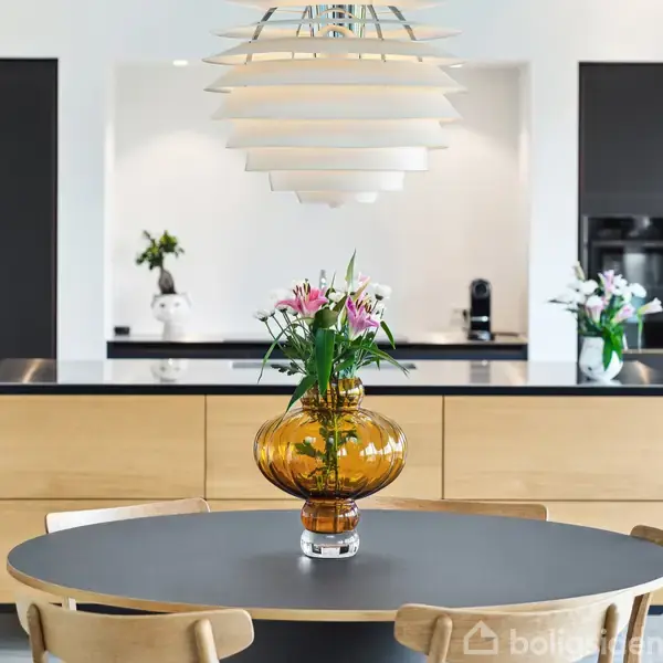 En rund bordplade med en brun vase fyldt med blomster står i centrum. I baggrunden er et moderne køkken med sorte og træfarvede skabe. Lysekrone over bordet.