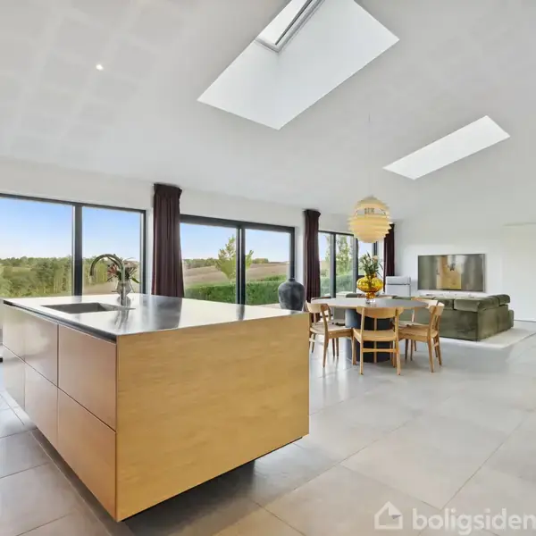 Et moderne køkkenalrum med en stor køkkenø, hvorpå der står en plante. Ved siden af er der et spisebord med stole og store vinduer med udsigt til natur.