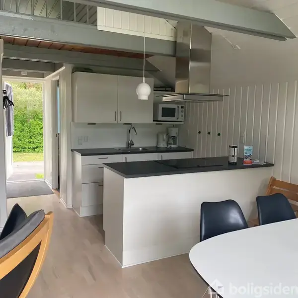 Køkken med hvide skabe og sort bordplade, indeholder mikroovn, kaffemaskine og emhætte. En åben dør fører til en have med grønne buske. Rundt om et bord står stole.
