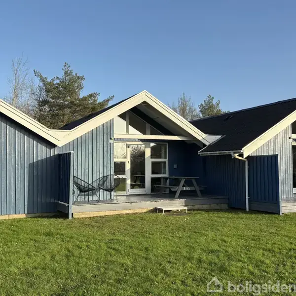 Blåtræbeklædt hus med store vinduespartier og trekantede gavle. Foran huset står et par stole og et havebord på en terrasse. Omgivet af græsplæne og træer i baggrunden.