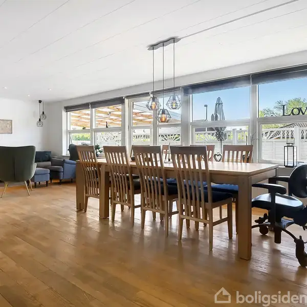 En spisestue med et langt spisebord og mange stole. Store vinduer lader lys komme ind, og ordene "Love" er placeret på en hylde. Der ses en sofa og lænestol i baggrunden.