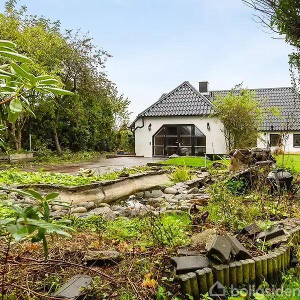 Huset har hvid pudset facade og sort tegltag, står i en frodig have med en lille dam omgivet af sten og beplantning, der er omgivet af forskellige træer og buske.