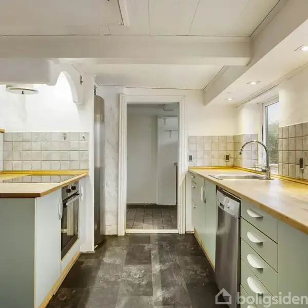 Køkken med lysegrønne skabe og træbordplade, indeholder indbygningsovn og opvaskemaskine. Mosaikfliser på væggen. Rummet er lyst med indbygget belysning og har en dør til et andet rum.