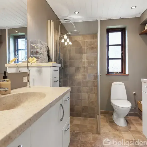 Badeværelse med håndvask og spejl, rummeligt bruseområde med glasvægge, og toilet ved siden af vindue. Hylder med dekorative genstande skaber en indbydende og moderne atmosfære.