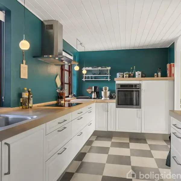 Et lyst køkken med hvide skabe og gråt ternet gulv. En vask og ovn ses langs væggen. Blå vægge skaber kontrast i det solbeskinnede rum, uden synlig tekst.