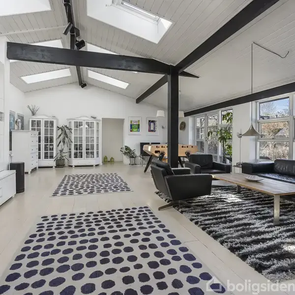 Stue med mørke bjælker under loftet, læder hjørnesofa og lænestol på mønstrede tæpper. Store vinduer giver lys til rummet, der er dekoreret med hvide møbler og grønne planter.